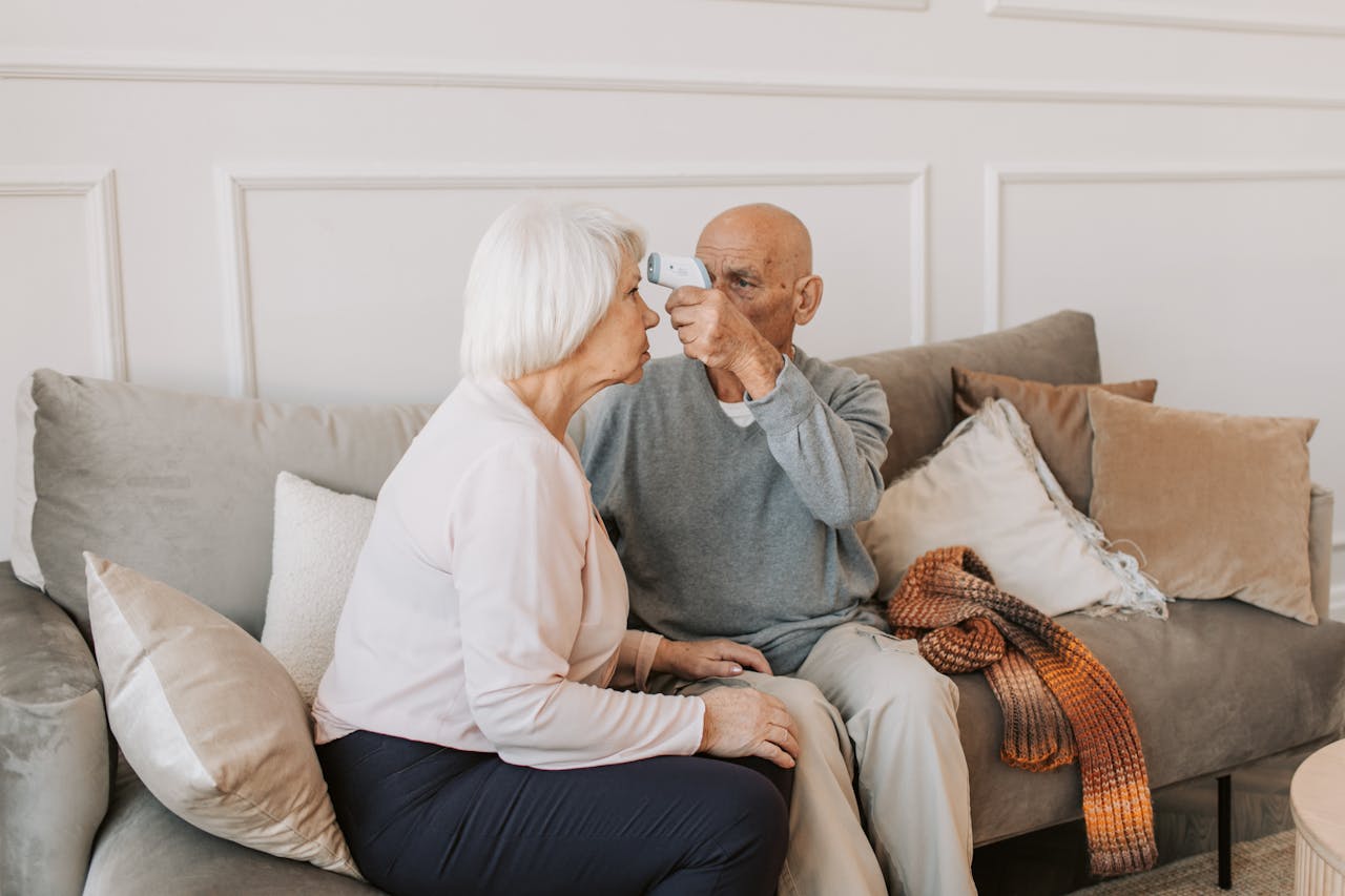 Elderly couple sitting on a sofa, checking temperature indoors, symbolizing togetherness and care.